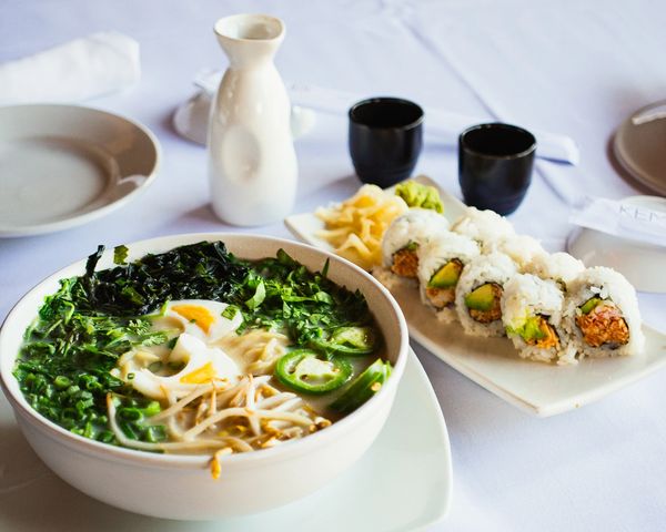 A bowl of ramen with boiled egg and greens, alongside a plate of sushi rolls with avocado and spicy filling.