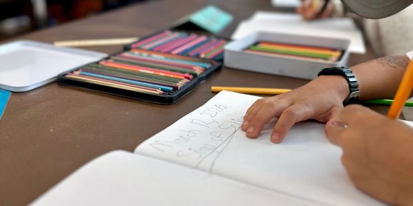 A child writes in a notebook with colored pencils nearby.
