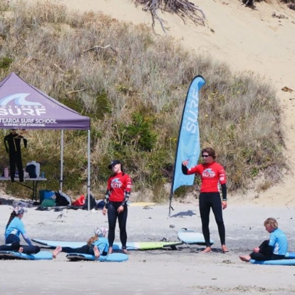 Surf instructors teaching kids on the beach near a surf school tent and banner.