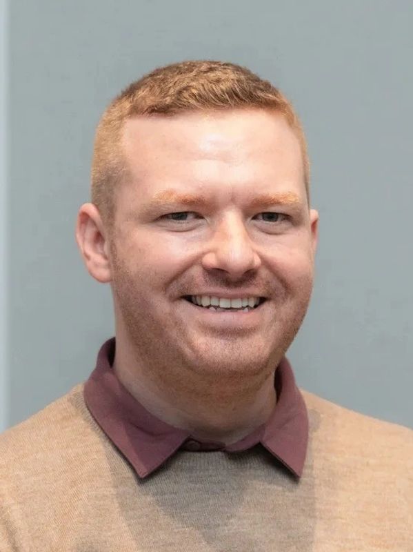 Smiling man with short red hair wearing a beige sweater and maroon collared shirt.
