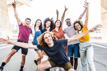 Group of diverse young friends joyfully posing outdoors with arms raised.