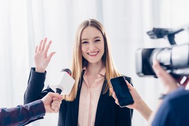 Smiling woman waving during an interview with microphones and a camera.