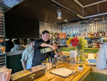 Bartender preparing a cocktail at a well-stocked bar with flowers on the counter.