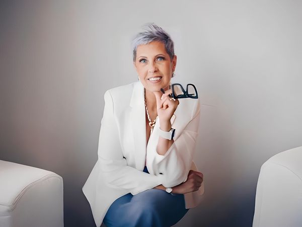A stylish person with short, light hair wears a white blazer, holding glasses