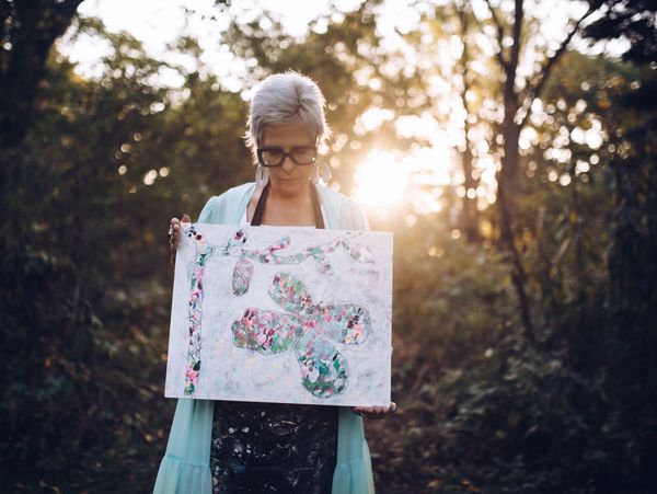 A person holds a colorful painting of a flower and vine in a forest setting