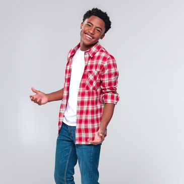 A person stands casually with arms open, wearing a red plaid shirt over a white t-shirt.