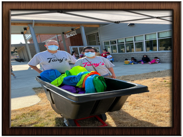 Two masked individuals pushing a cart with colorful bags outside a school.