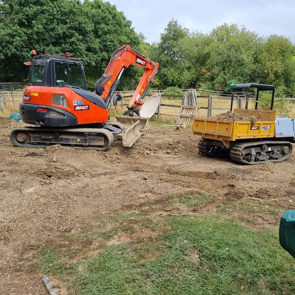 Two tracked construction vehicles working on a dirt area near a fence and trees.