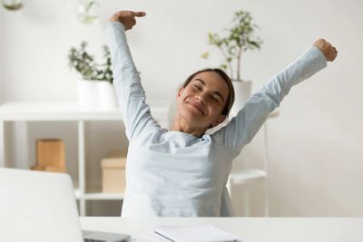 Young female sitting at desk hold arms in the air expressing triumph