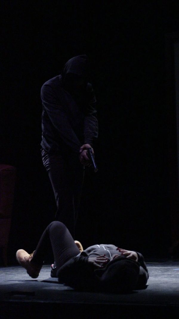 Someone on a stage is standing over a woman with a gun. They are dressed identically.