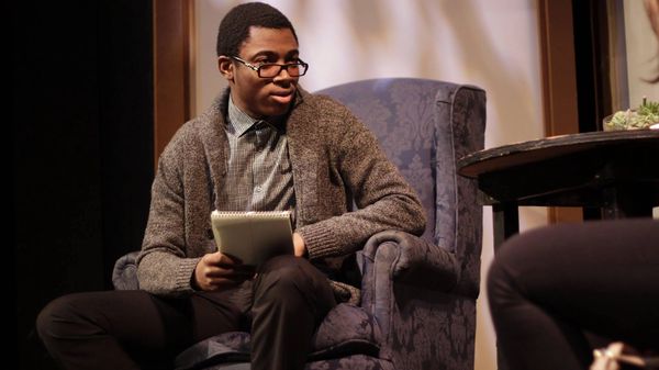 A male therapist wearing glasses and holding a note pad sits across from a woman slightly oo frame.
