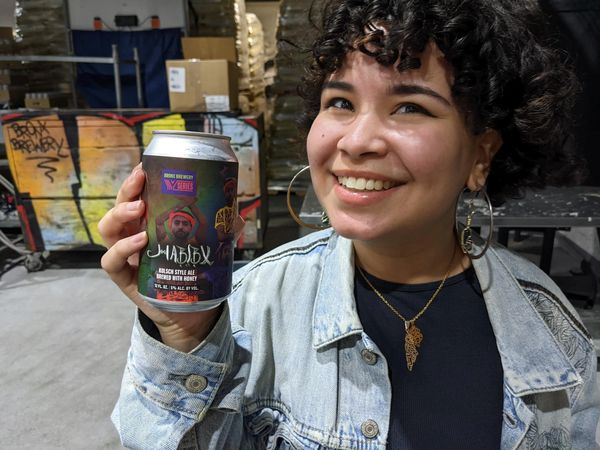 Lafi smiles holding a beer. They have curly short hair with bangs and hoop earrings.