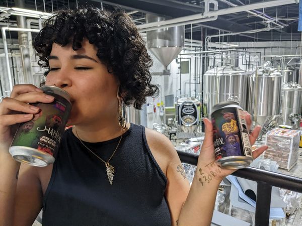 Lafi sips Habibx beer and holds another can in their other hand. Behind them i s the Bronx Brewery.