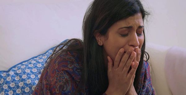 Woman is crying, holding her hands at her mouth. She has long hair and is sitting on a couch.