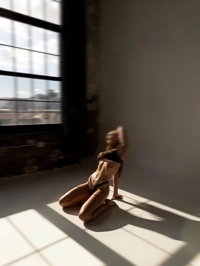 Silhouetted woman in black lingerie kneeling in sunlight by a large window.