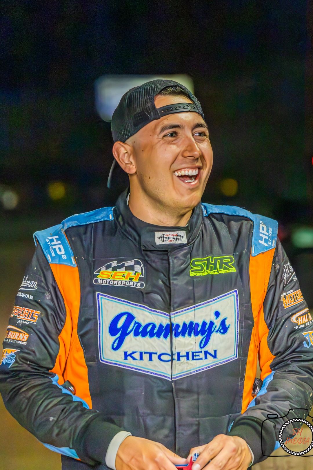 Smiling race car driver in a colorful suit and backward cap.