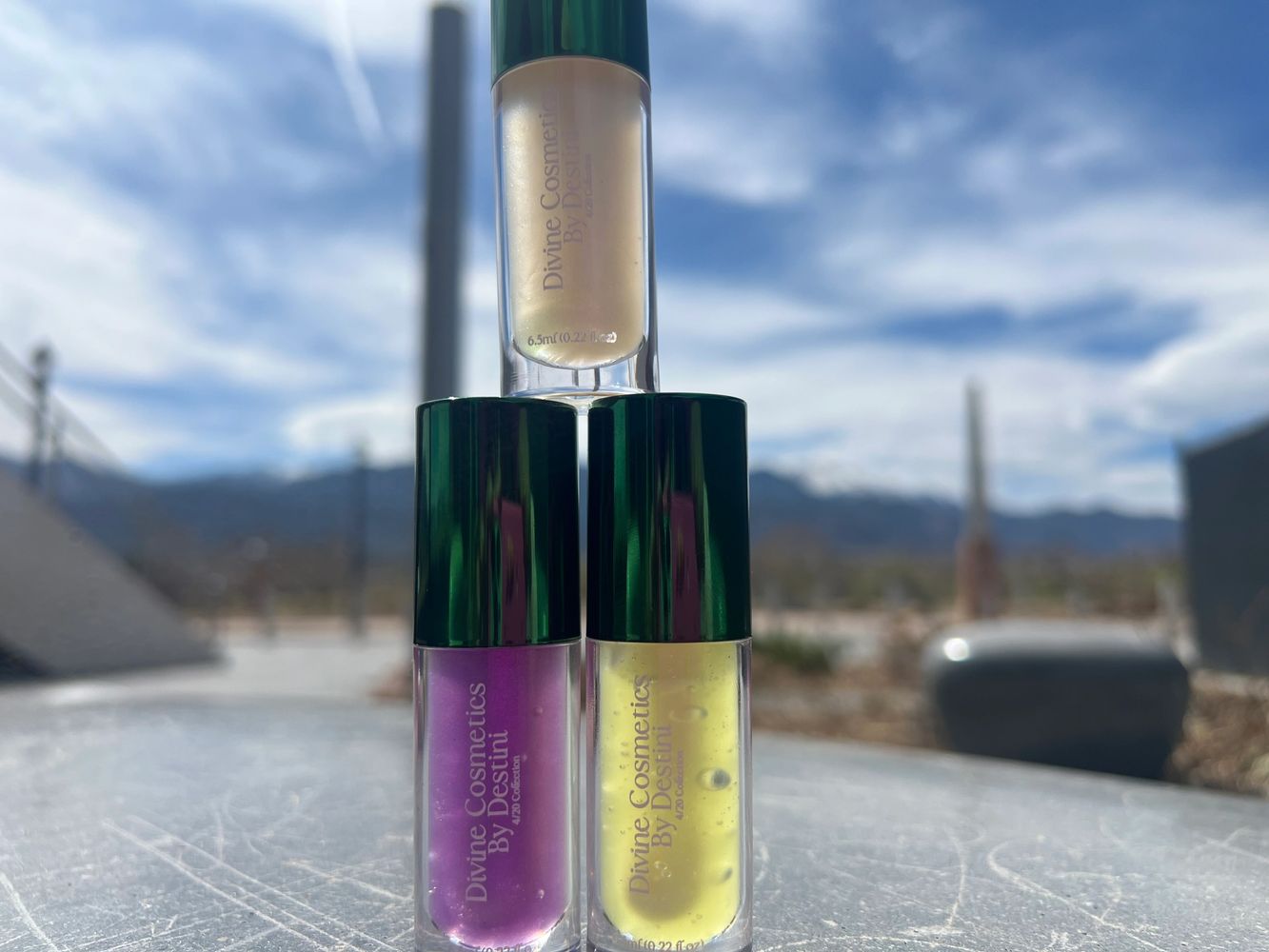 Three Divine Cosmetics lip glosses stacked outdoors under a cloudy sky.