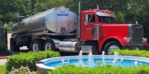 Red water tanker truck parked near a decorative garden fountain.