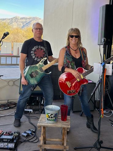 Two musicians posing with guitars next to a tip jar outdoors.