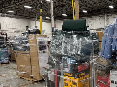 Liquidation pallets wrapped in plastic with various boxed appliances and furniture in a warehouse.