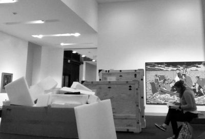 Rebecca in art gallery, sitting on bench, surrounded by art storage crates. 