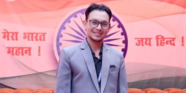 A man in a gray suit smiling with an Indian flag backdrop.