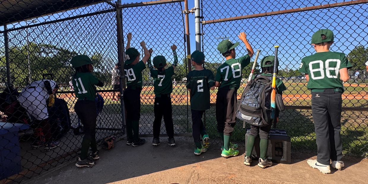 Young baseball players in green uniforms watch a game behind a chain-link fence.