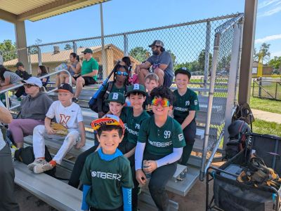 Youth baseball team sitting on bleachers under a shaded area.