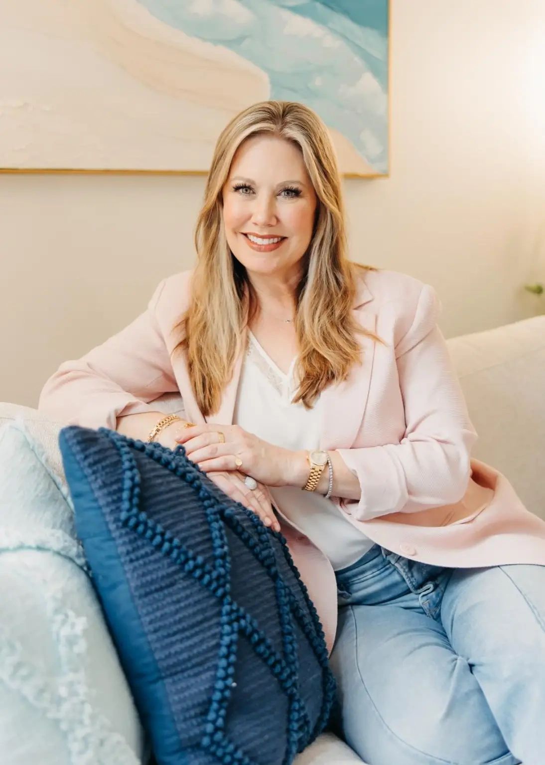 Smiling woman in a light pink blazer sitting on a couch with blue pillows.