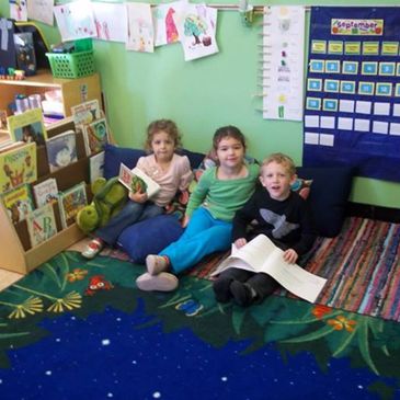 Three preschool students reading on classroom carpet and looking at camera for photo.