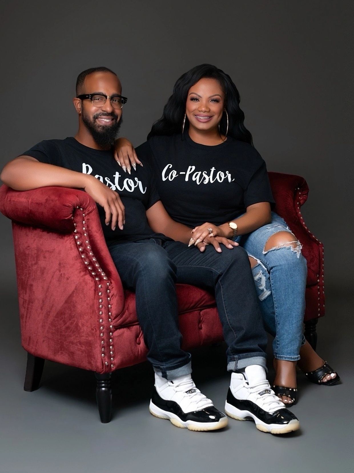 Couple wearing matching "Pastor" and "Co-Pastor" shirts sit on a red velvet couch smiling.