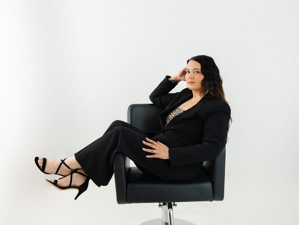 Woman in black suit sitting confidently on a black chair against a white background.