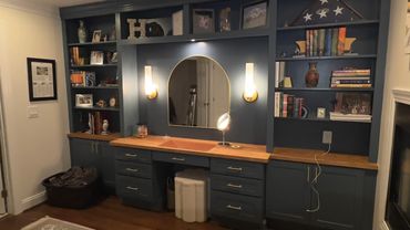 Cozy room with dark blue built-in shelves, wooden countertop, and a circular mirror with lights.