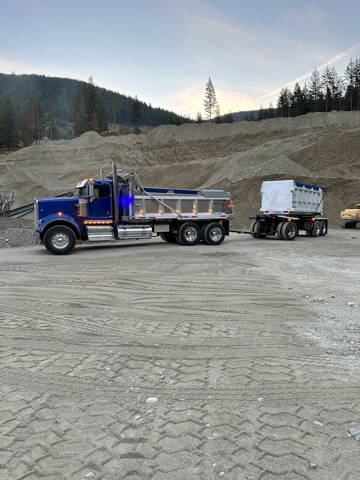A blue dump truck with a trailer parked at a gravel site near forested hills.