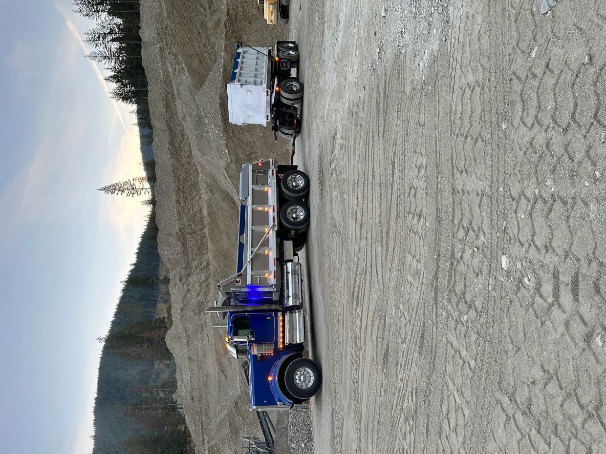 A blue dump truck with a trailer parked at a gravel site near forested hills.