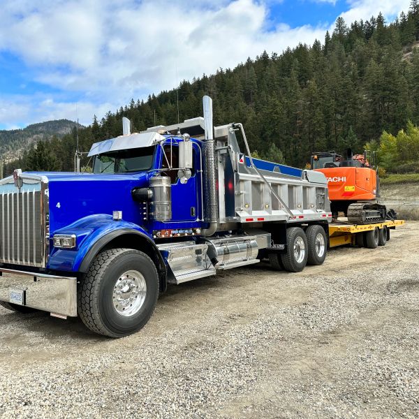 Blue dump truck parked on a gravel road with forested hills in the background.