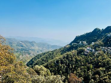 Mountain landscape with houses scattered on forested hills under a clear blue sky.