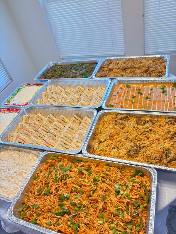 A spread of diverse dishes including sandwiches, noodles, and rice in large trays.