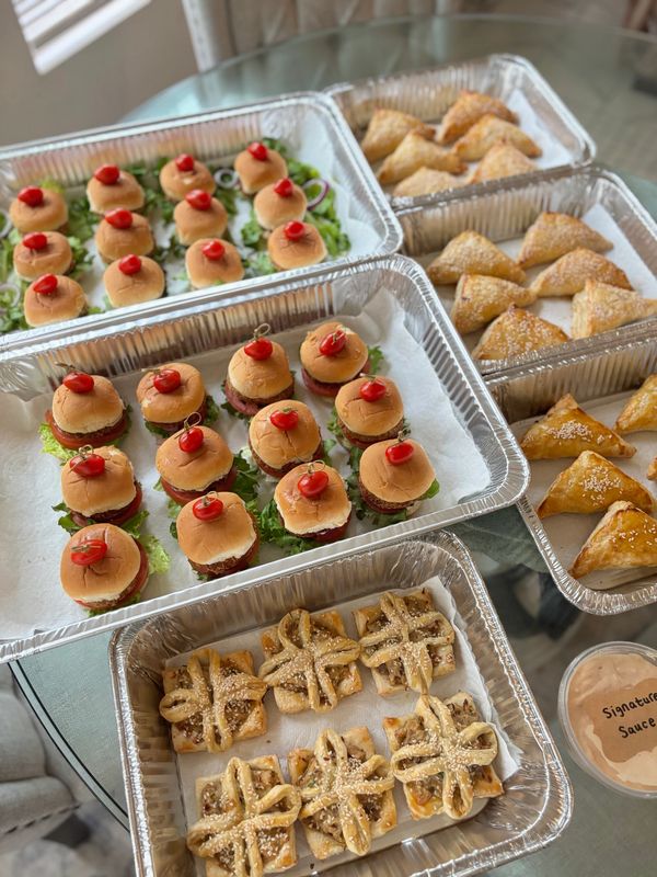 Trays of mini burgers, savory pastries, and signature sauce arranged on a glass table.