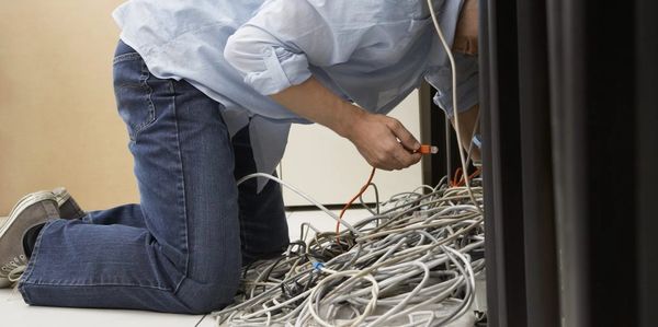 A person on their knees under a desk dealing with a mess of computer wires.