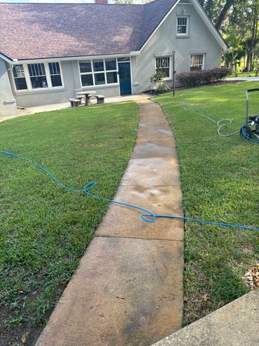 Concrete pathway leading to a house with a blue door, surrounded by green grass and garden furniture.
