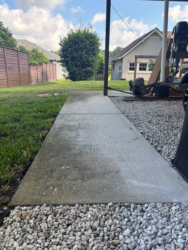 Concrete sidewalk bordered by grass and gravel leading to a backyard with houses.