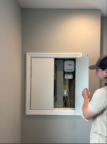 Man opening a utility panel door on a gray wall.