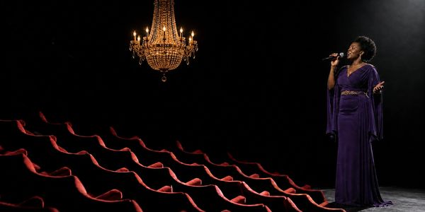 Female vocalist in purple singing on stage beneath one gold chandelier, with red auditorium seats.