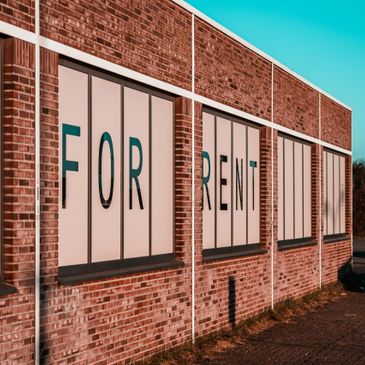 Brick building with large "FOR RENT" signs on windows under clear blue sky.