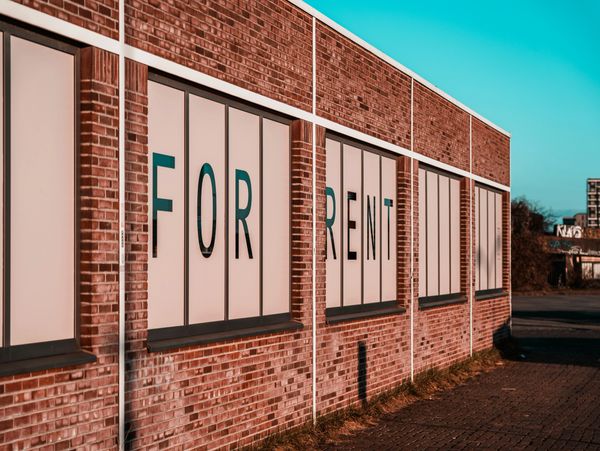 A brick building with large windows displaying a 'FOR RENT' sign.