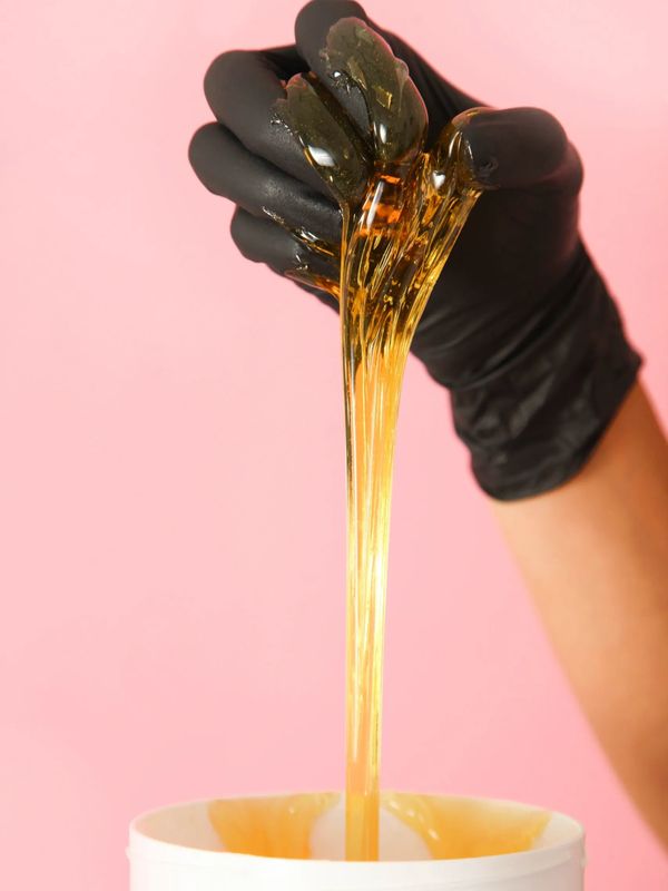 Black-gloved hand stretching golden honey-like wax over a pink background.