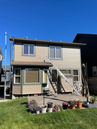 Backyard of a two-story house with a ladder and flower pots on a sunny day.