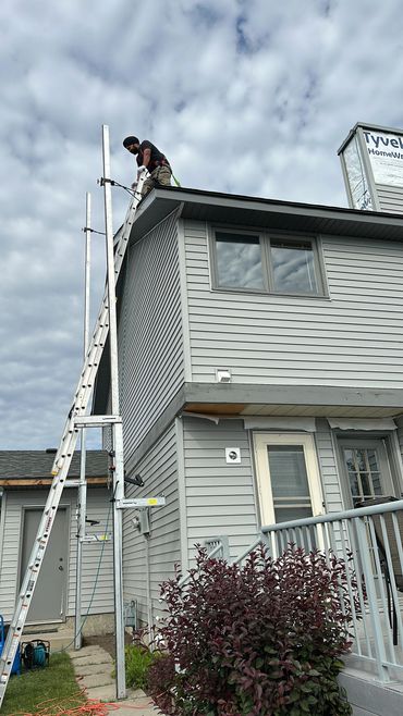 A man working on a ladder on the roof of a gray house under a cloudy sky.