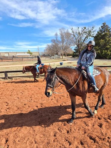 Two people enjoying horseback riding in a sunny outdoor arena.